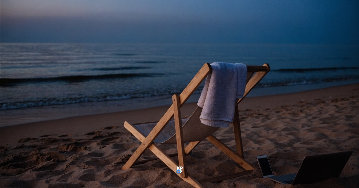 Leerer Liegestuhl mit Handtuch am Strand in blauer Abenddämmerung, im Sand daneben ein aufgeklapptes Notebook und ein Smartphone, im Hintergrund das Meer