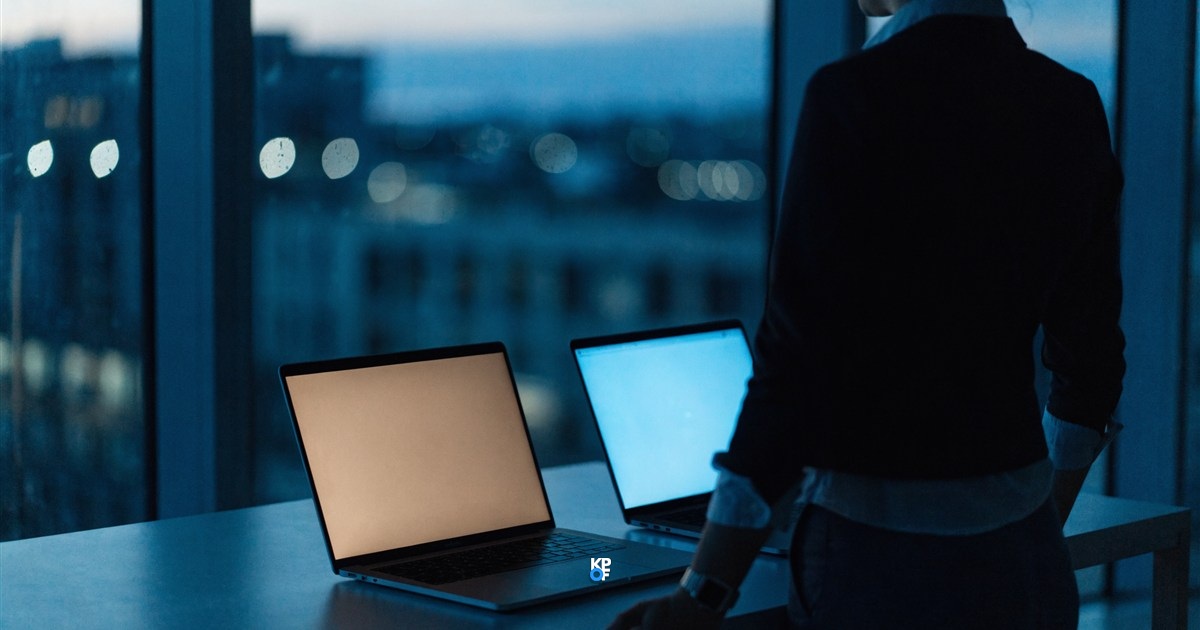 Zwei Laptops einander zugewandt auf einem Schreibtisch am Abend, ein Bildschirm leuchtet warm, der andere kühl blau, daneben eine Person vor der Fensterfront mit Blick auf die Stadt
